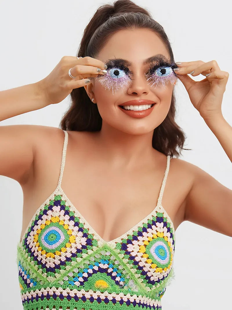 Smiling woman wearing colorful crochet top holding handmade surrealist eye earrings in front of her eyes