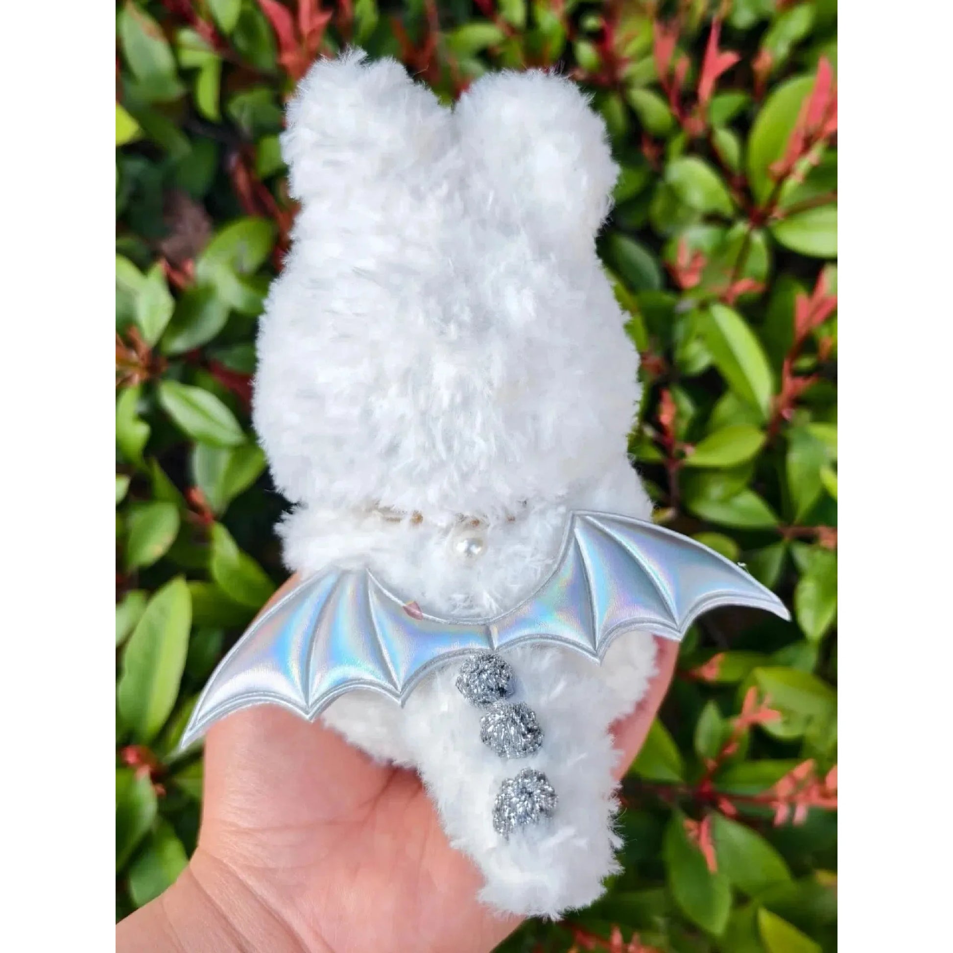 Hand holding a fluffy white crochet figure with shiny silver wings and three pom pom buttons angel costume crochet pattern
