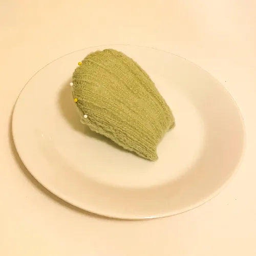Green knitted madeleine dessert pattern displayed on a white plate with pins attached